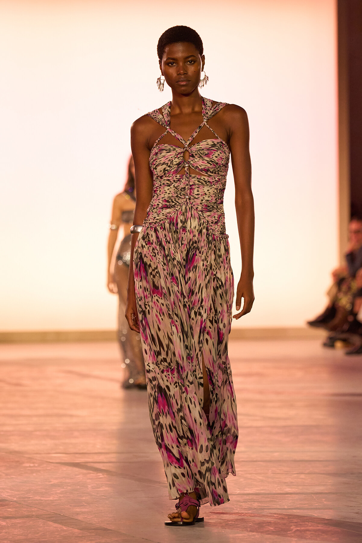 Model walks a fashion runway in a pink‑purple abstract print halter dress with cutouts and crisscross straps, pink sandals.