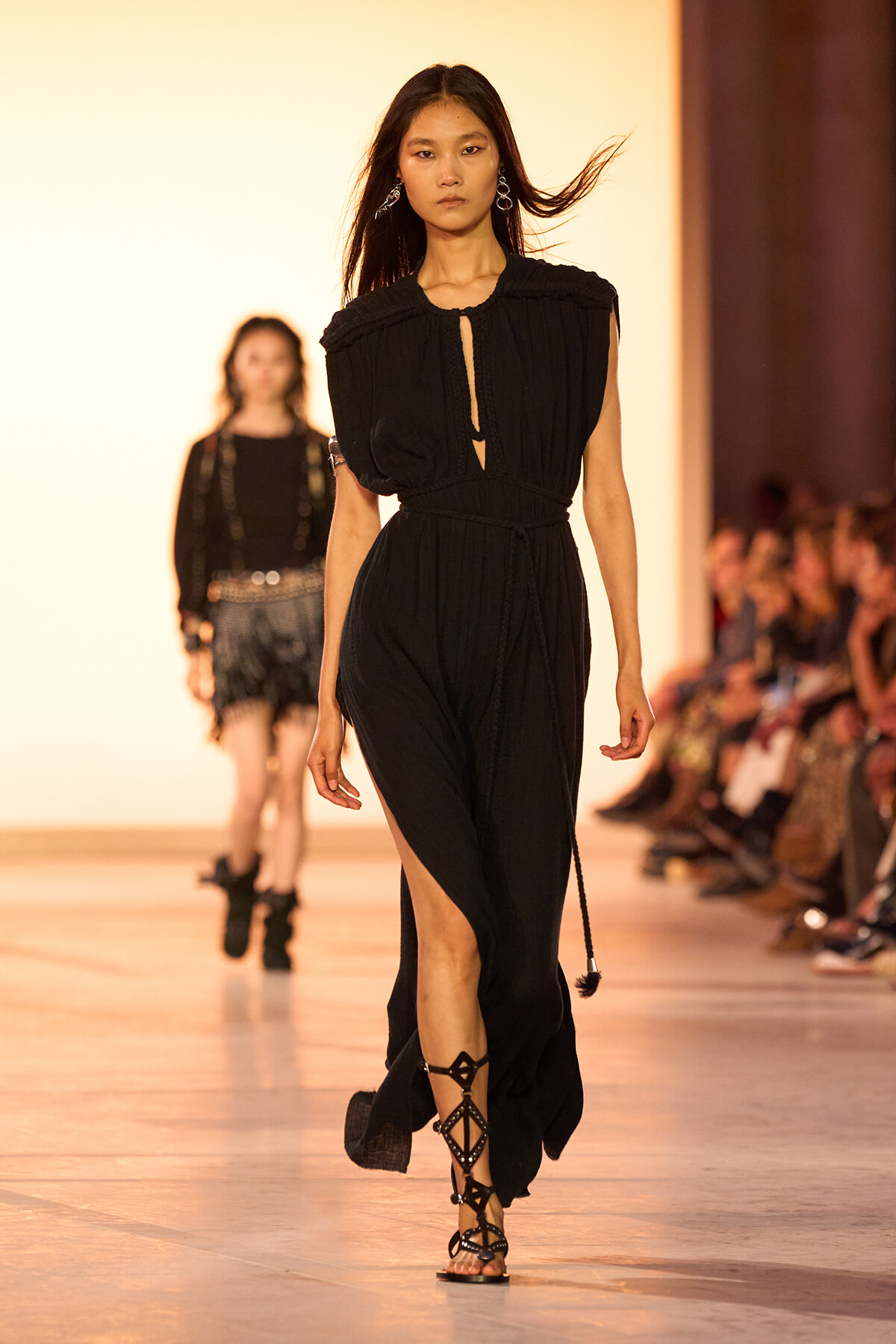 Model walking the runway in a black sleeveless dress with a waist belt and high side slit; audience visible on the right side of the catwalk.