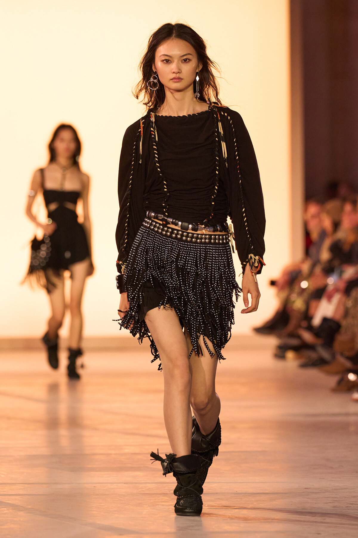 Model walks a runway in a black fringed mini skirt, ruched top, and layered straps with metallic accessories.