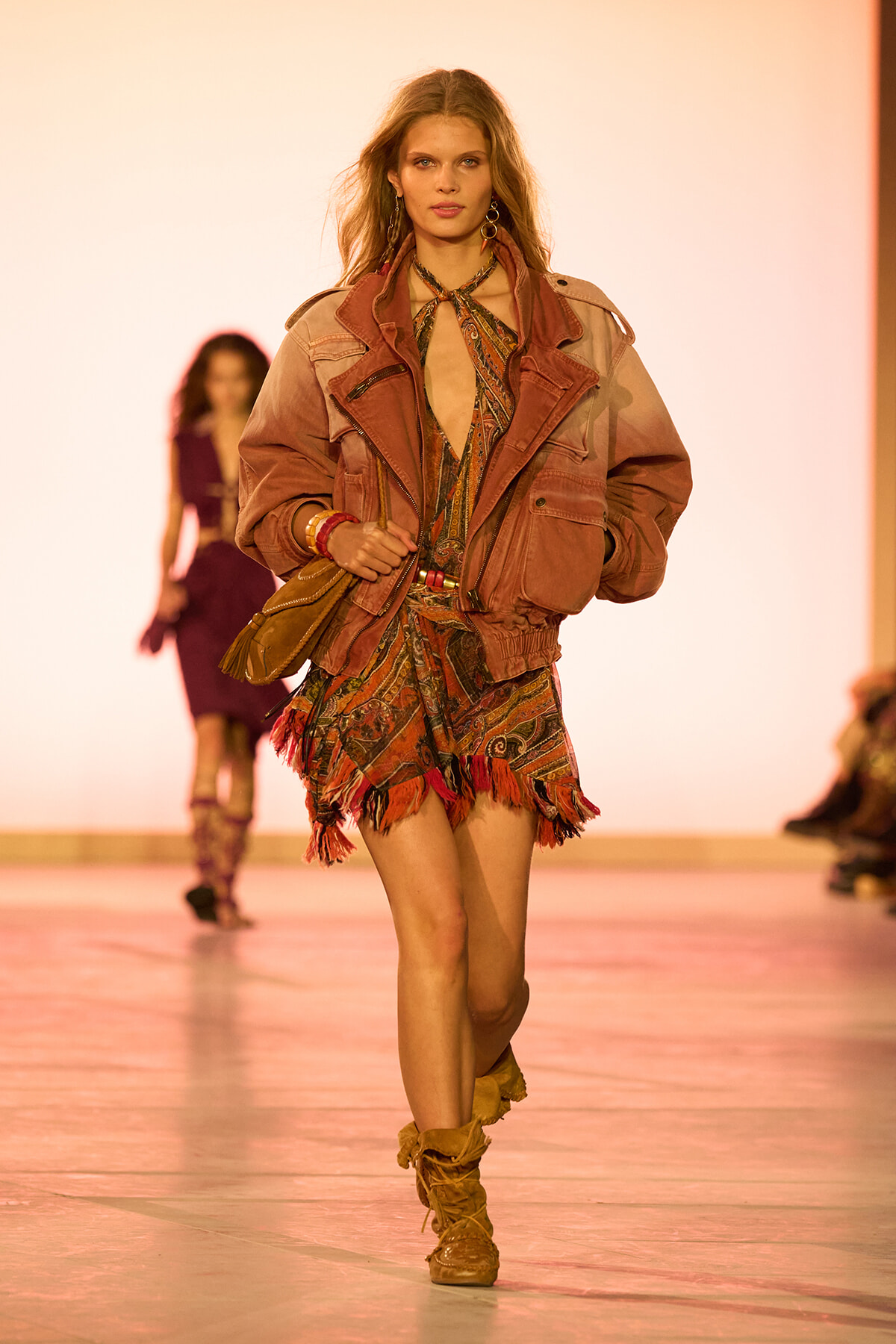 Model walking runway in a dusty pink oversized jacket over a patterned printed romper with fringe hems, tan boots, and boho scarf at neck.