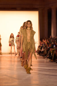 Model walks a runway in a beige fringed outfit with knee-high boots, during a fashion show.