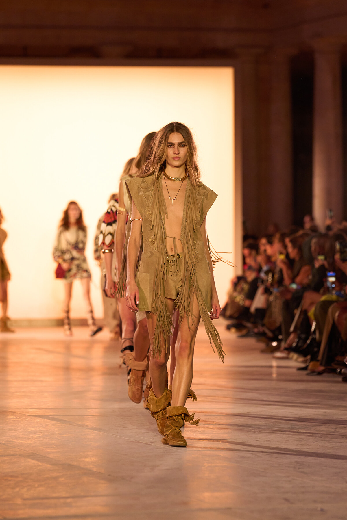 Model walks a runway in a beige fringed outfit with knee-high boots, during a fashion show.