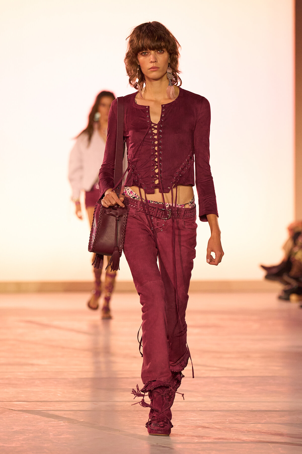 Model walking the runway in a burgundy laced crop top and matching low-rise pants with fringe boots and a shoulder bag.