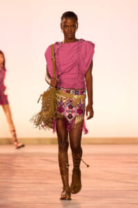 Model walking a fashion runway in a pink ruched top, a colorful fringed mini skirt, knee-strap sandals, and a tan fringed shoulder bag.