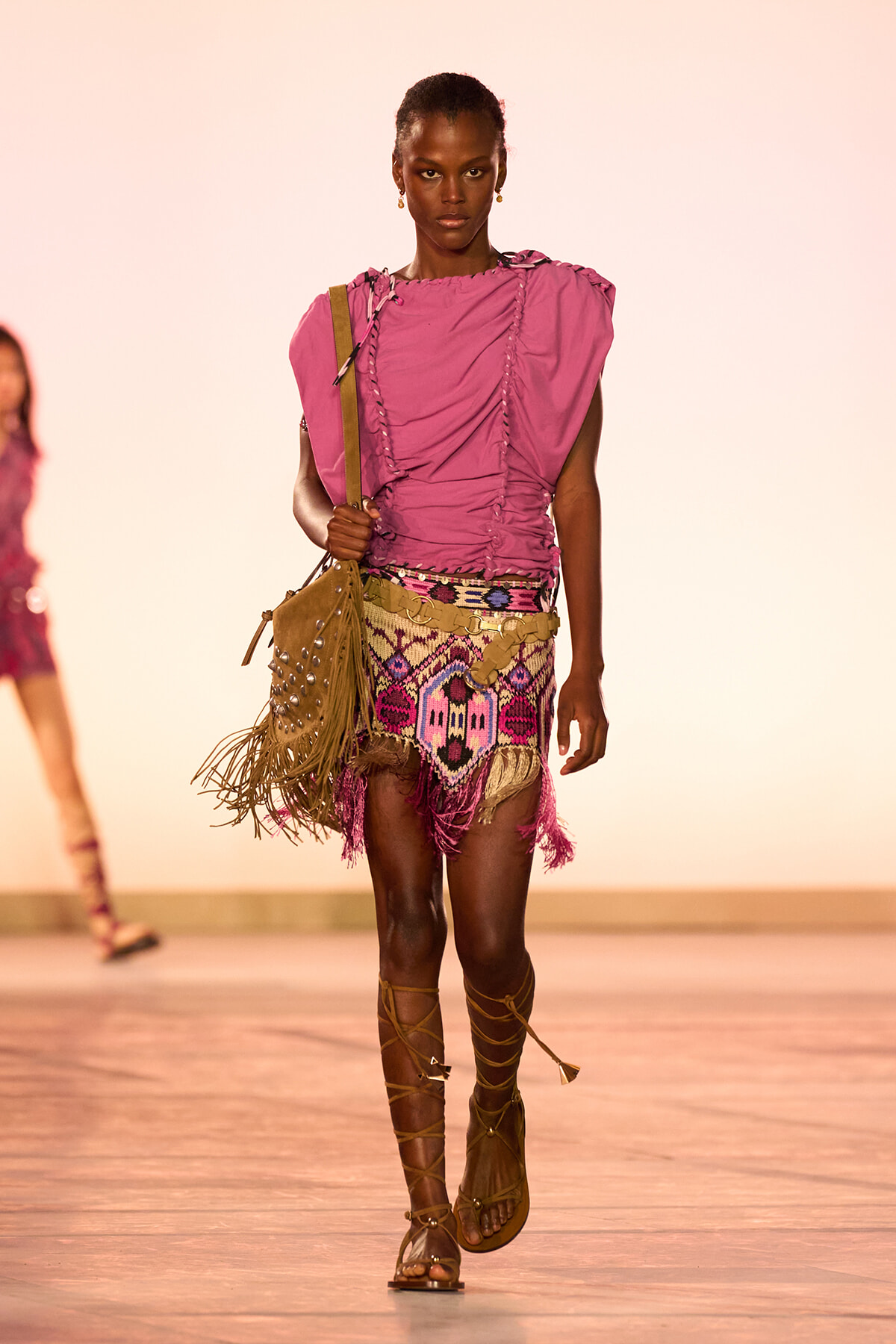 Model walking a fashion runway in a pink ruched top, a colorful fringed mini skirt, knee-strap sandals, and a tan fringed shoulder bag.
