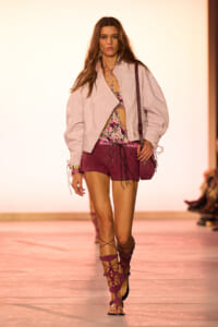Model walking a runway in a pale pink oversized jacket, pink floral top, burgundy shorts, and strappy burgundy sandals with a small bag equipped.