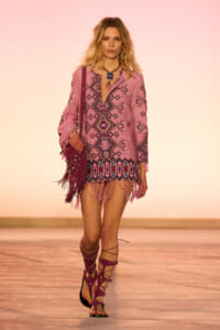 Model walking a runway in a pink, embroidered boho tunic with fringe at the hem, carrying a studded purple fringe bag and wearing strappy gladiator sandals.