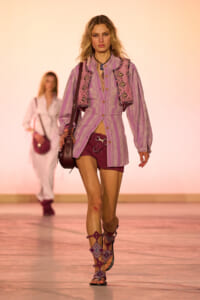 Model walks a fashion runway wearing a lavender striped shirt-dress with a pink embroidered vest, burgundy shorts, gladiator sandals, and a crossbody bag visible against a peach backdrop.