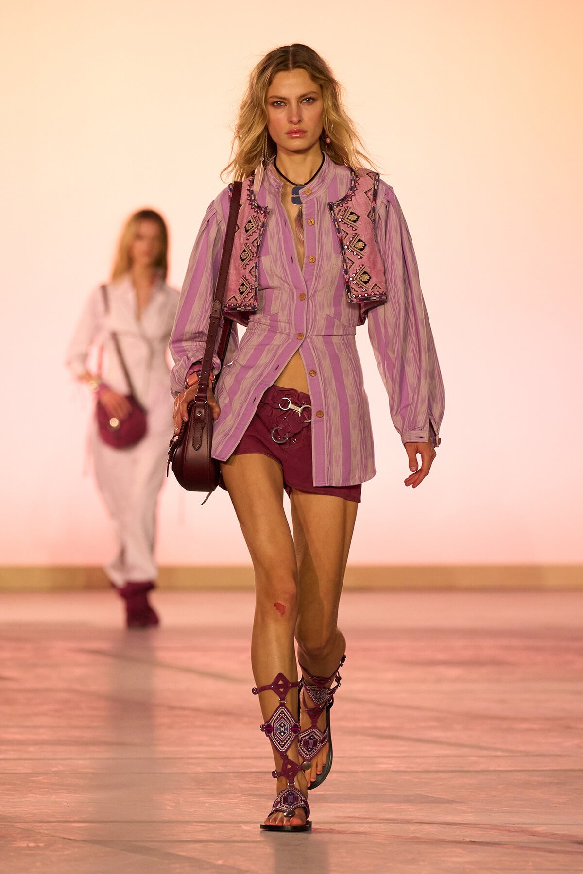 Model walks a fashion runway wearing a lavender striped shirt-dress with a pink embroidered vest, burgundy shorts, gladiator sandals, and a crossbody bag visible against a peach backdrop.