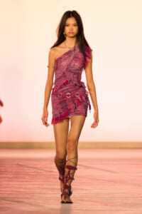 Runway model wearing a pink one-shoulder fringed mini dress with a silver ring belt and gladiator sandals.