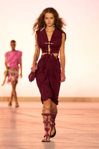 Model walking a runway in a burgundy cut-out jumpsuit with knotted detailing, paired with gladiator sandals and a small burgundy bag.