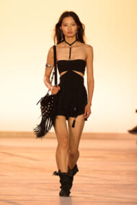 Female model walking a fashion show runway in a black strapless top with cutouts, high-waisted shorts, and a fringe-studded bag with black boots