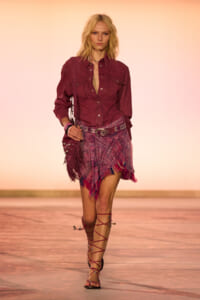 Model walking the runway in a burgundy button-up shirt, pink patterned fringed wrap skirt, belt, crossbody fringe bag, and strappy gladiator sandals.