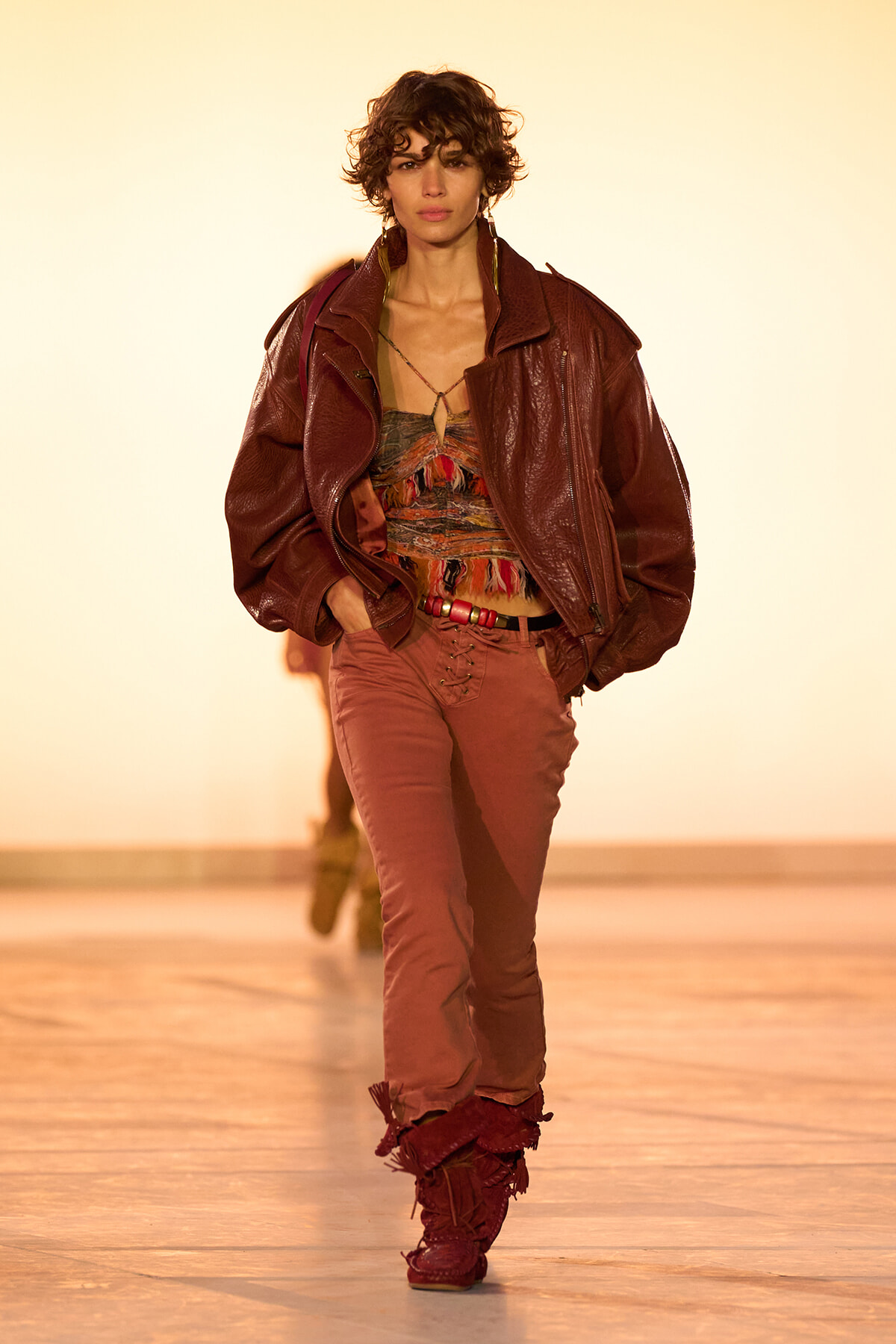 Runway model wearing a oversized burgundy leather jacket, patterned top, pink pants, and fringed boots walking the catwalk.