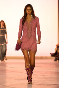 Model on a runway wearing a pink lace-up tunic dress with scalloped hem, matching pink gladiator sandals and purse.