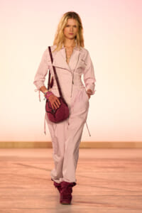 Model walking a runway wearing a light pink jumpsuit with a front zipper, burgundy crossbody bag, and burgundy boots, accessorized with bracelets.