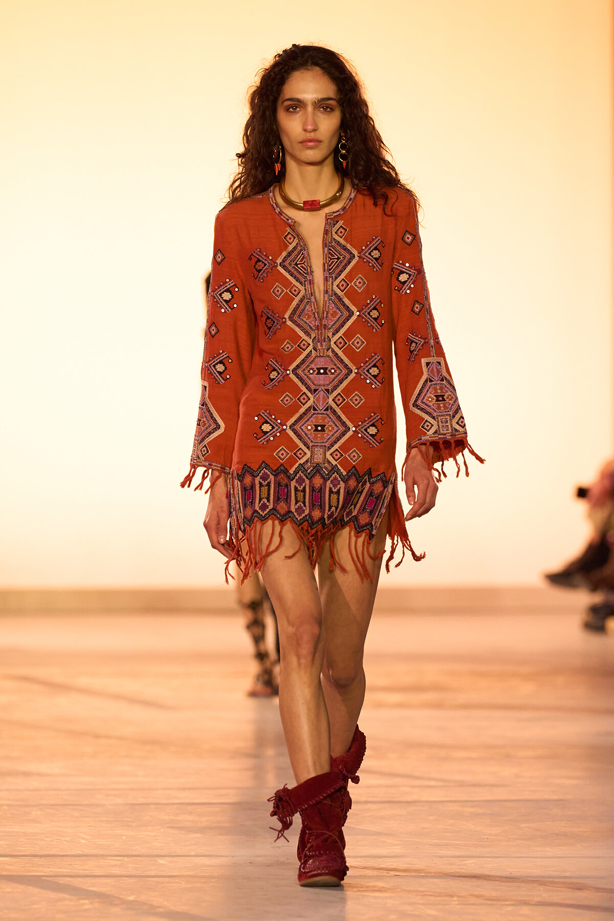 Model walking a runway in an orange bohemian tunic with geometric embroidery and fringe, paired with red boots.