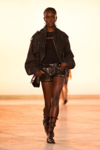 Model walks the runway in an all-black look: oversized jacket, studded shorts, and leather gladiator sandals with a small bag.