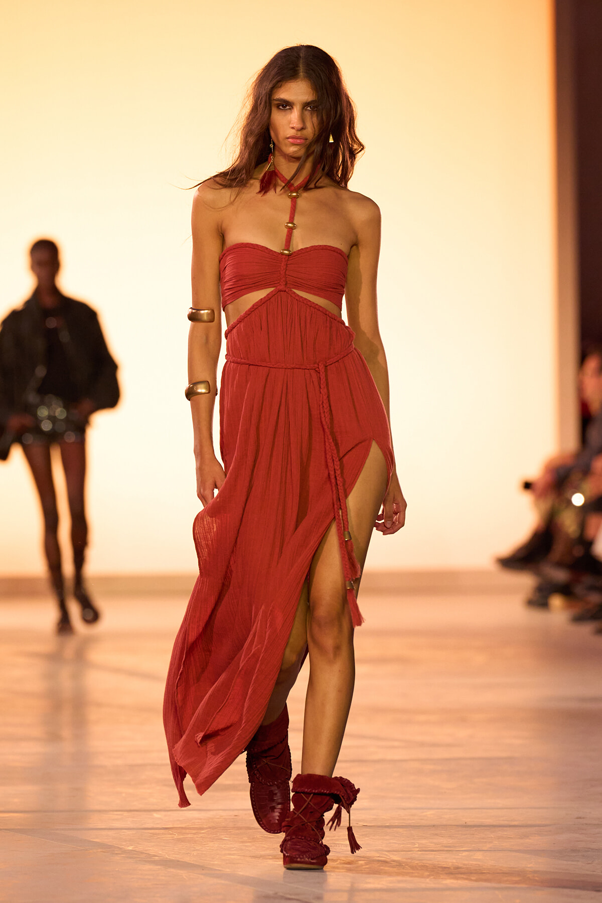 Model walking the runway in a red, strapless dress with a high slit and flowing skirt, during a fashion show.