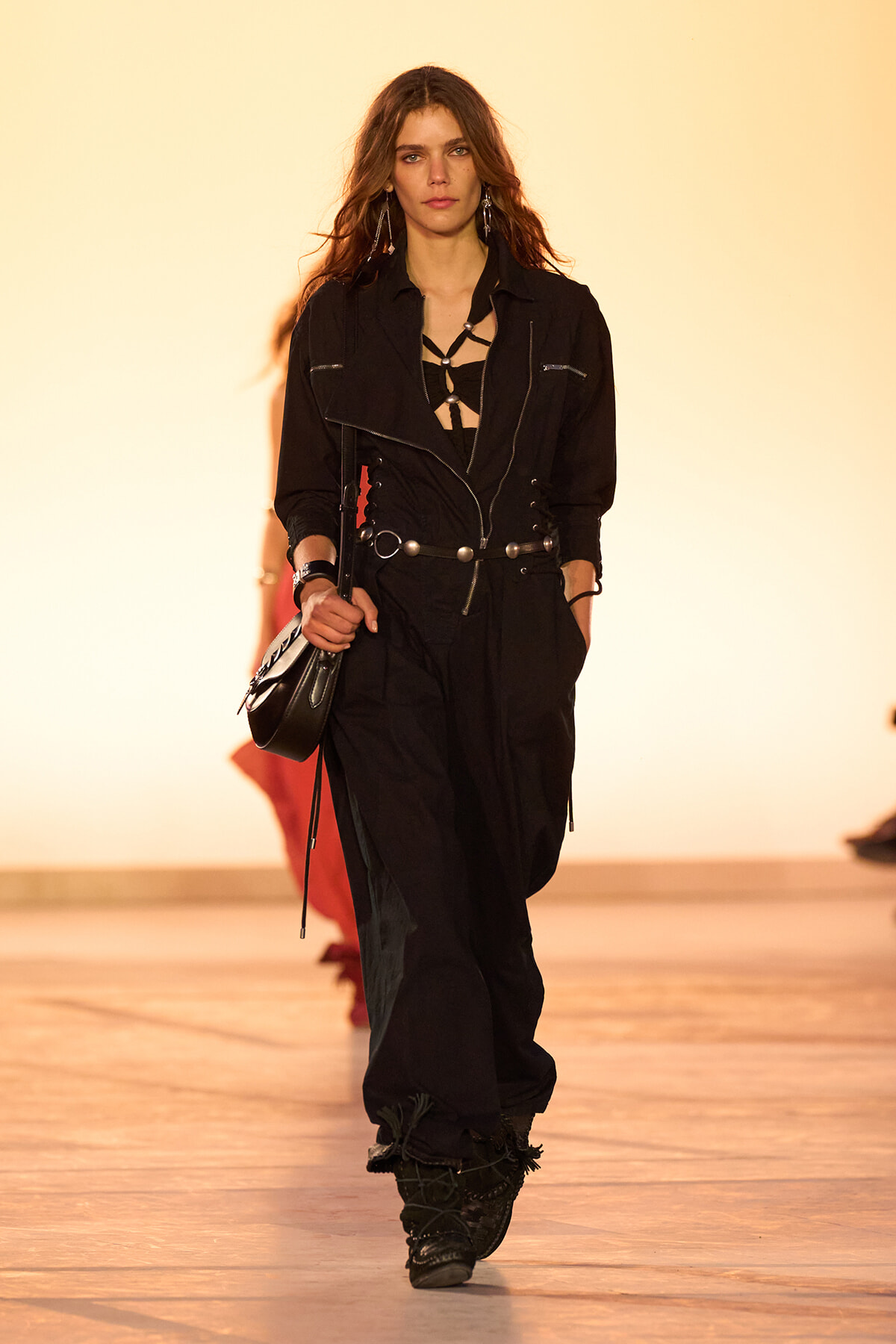 Female model walks a runway in an all-black outfit with a harness-style top, wide belt with silver studs, and a small black bag.