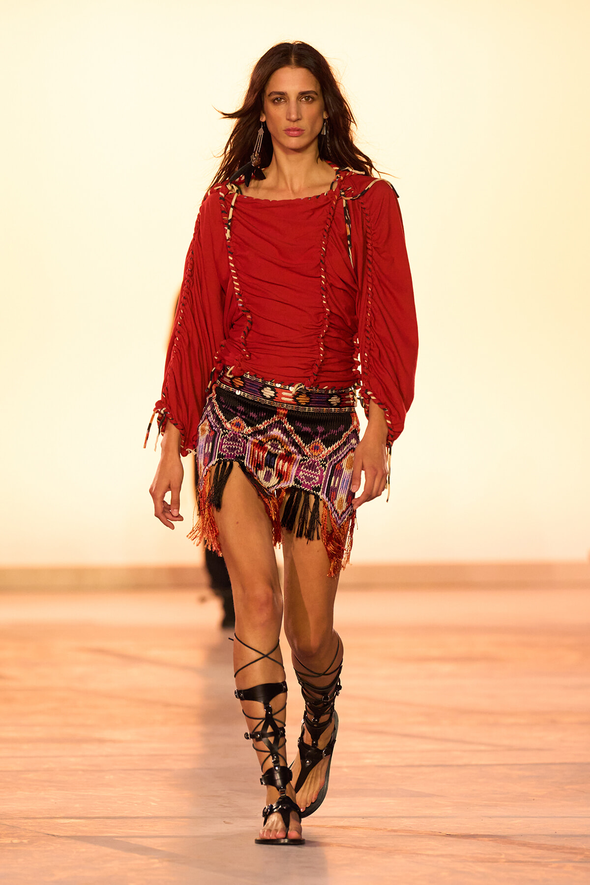 Model walking runway in a red ruched blouse with braided trim and a multicolor fringe mini skirt with tassels, black gladiator sandals.