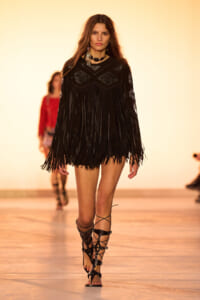 Model walks the runway in a black fringed dress with geometric chest panels and strappy gladiator sandals on a fashion show catwalk.