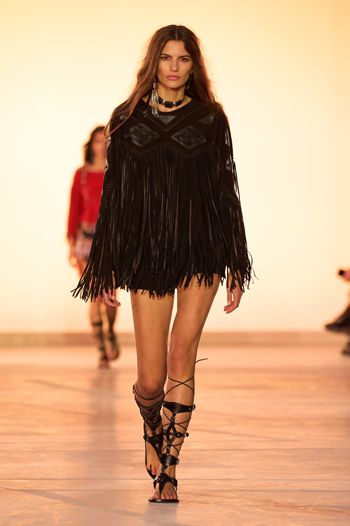 Model walks the runway in a black fringed dress with geometric chest panels and strappy gladiator sandals on a fashion show catwalk.