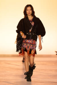 Model walking a fashion show runway in a black oversized jacket over a colorful patterned fringe dress with fringe details and black boots, carrying a studded black bag.