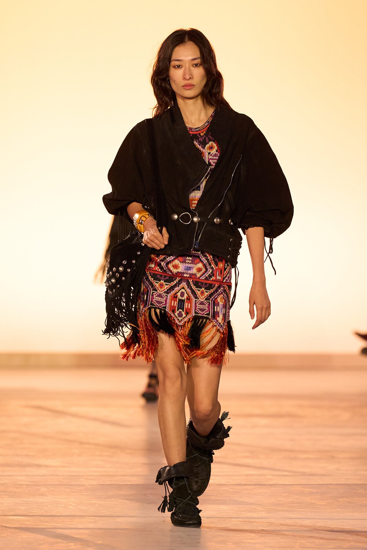 Model walking a fashion show runway in a black oversized jacket over a colorful patterned fringe dress with fringe details and black boots, carrying a studded black bag.