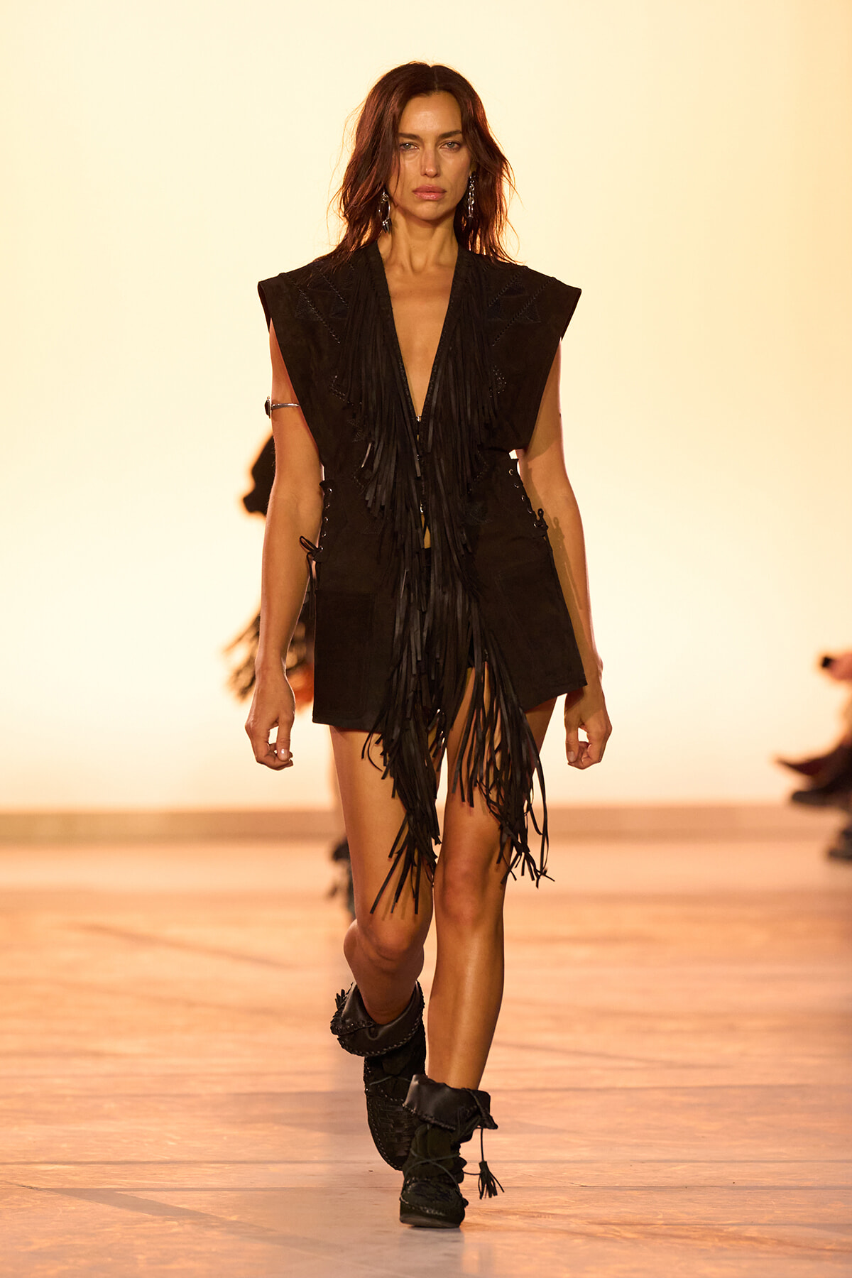 Model walks the runway in a black fringe-vest outfit with shorts and black boots, against a warm yellow backdrop.