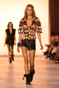 Model walking the runway in a bold geometric-patterned top with a scoop neckline and black studded shorts, accessorized with black boots.