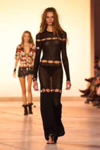 Model on a runway wearing a sheer black knit jumpsuit with circular cut-out bands at the chest, waist, and legs, paired with sandals.