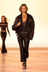 Model walking the runway in a black leather jacket, ripped-front top, and matching leather pants under warm stage lights.