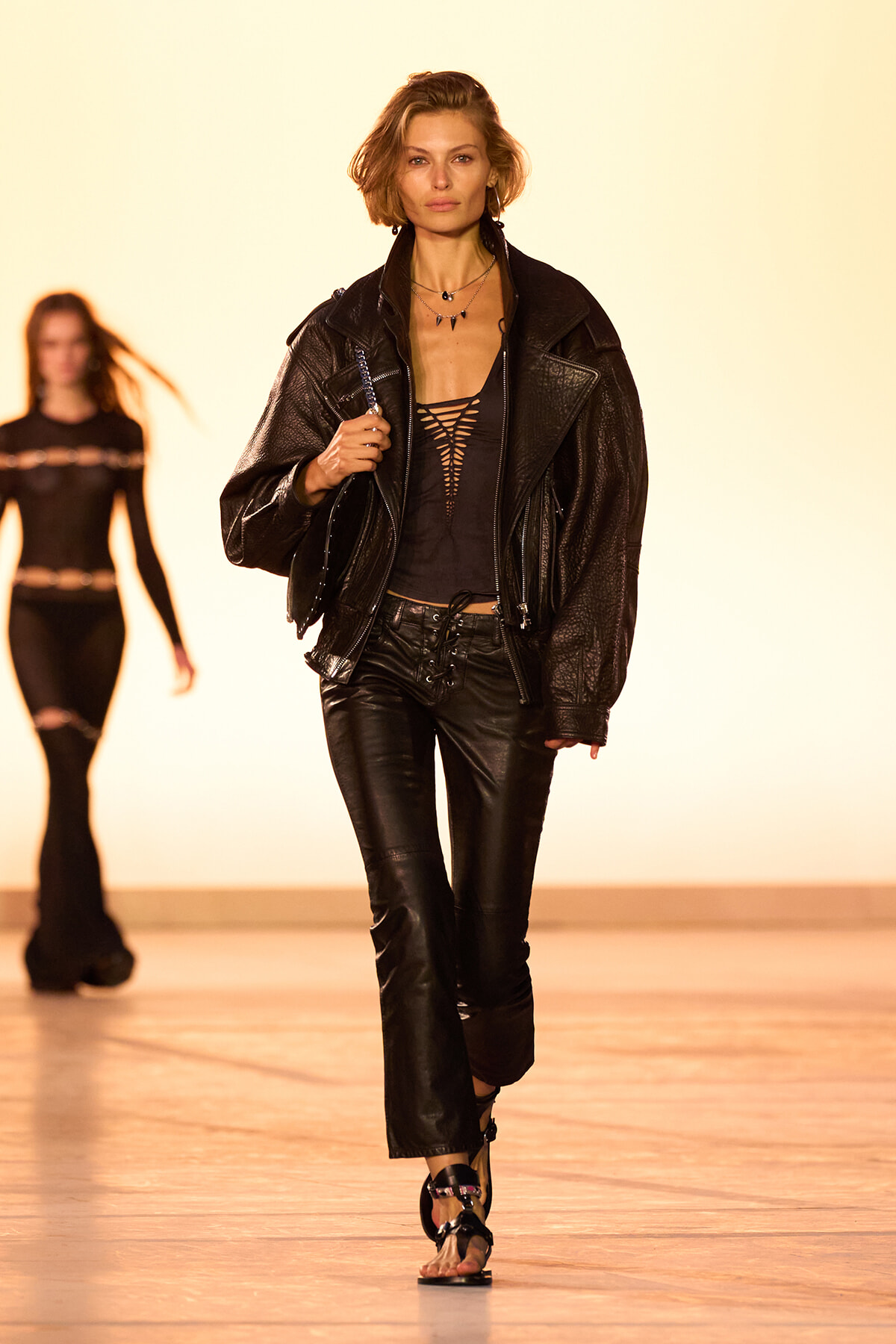 Model walking the runway in a black leather jacket, ripped-front top, and matching leather pants under warm stage lights.