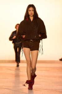 Model walking a runway in a dark brown oversized top, brown shorts, and red ankle boots with red accessories