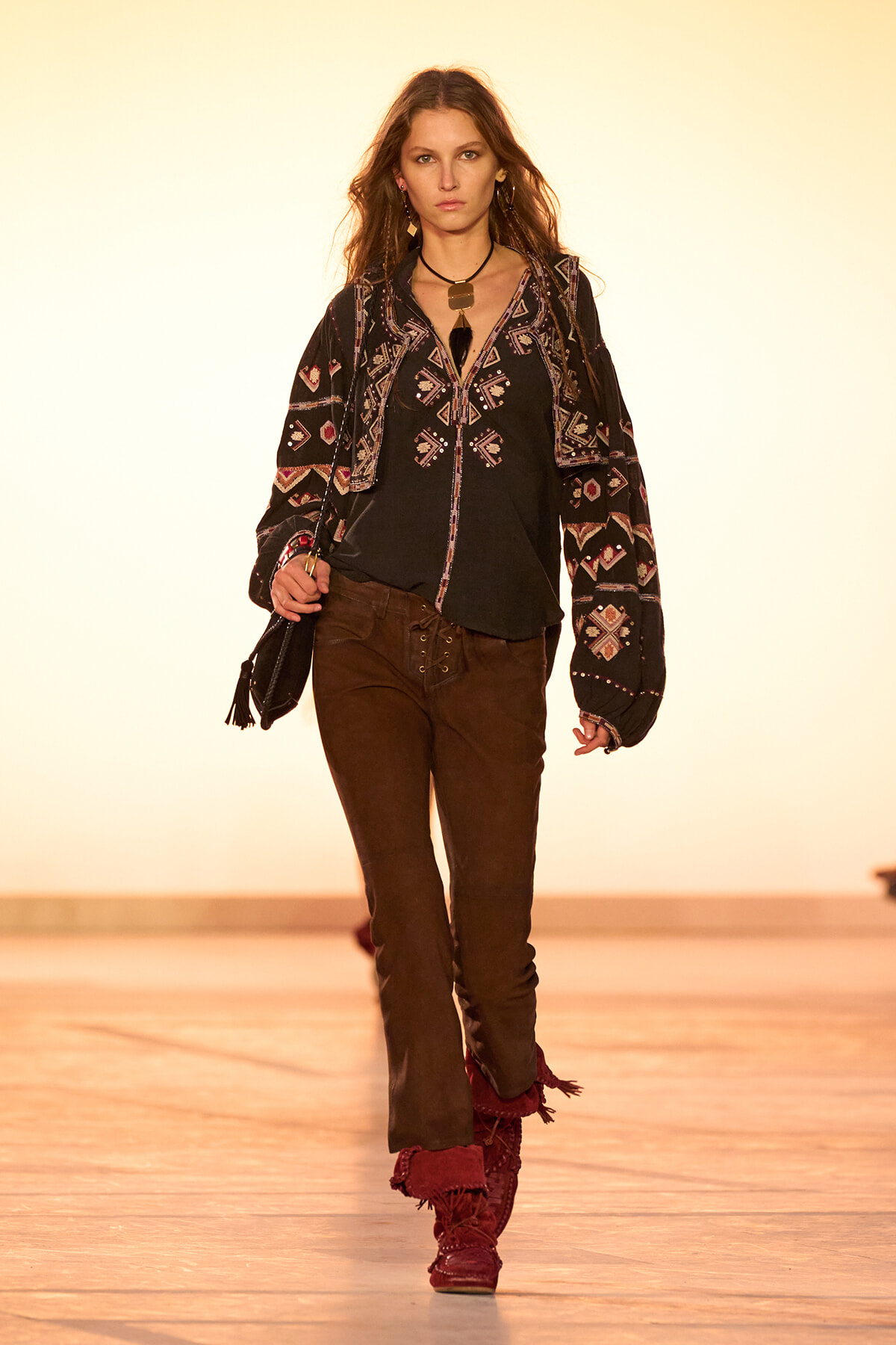 Model walking runway in a black embroidered blouse, brown pants, and red fringed boots, holding a small bag.