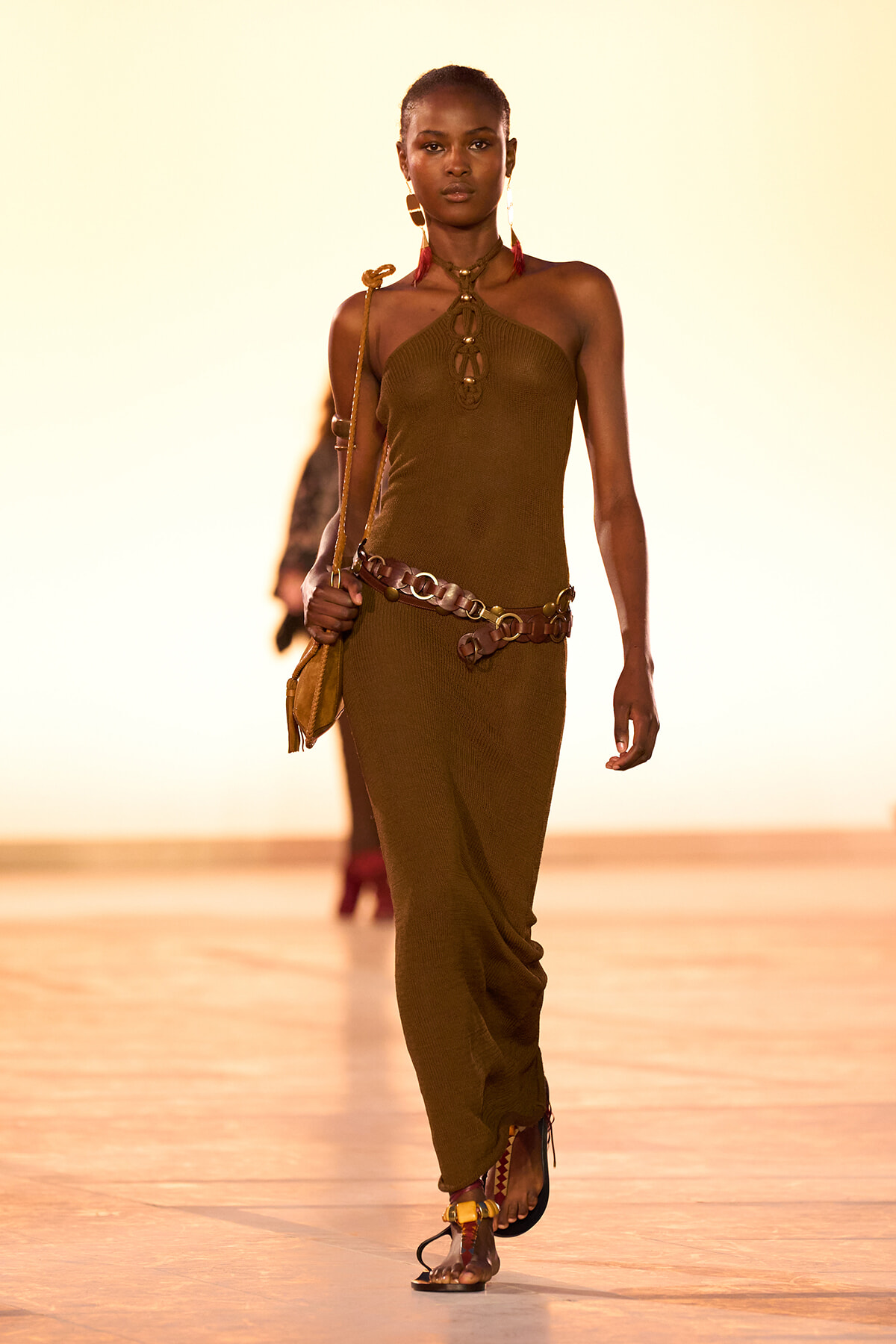 Model walking the runway in a brown halter-knit dress with a leather belt and small bag.