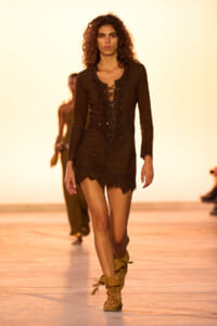 Model walks the runway in a dark brown lace-up mini dress with scalloped hem and tan fringe boots.