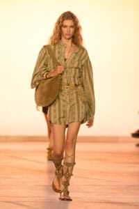 Model walking a runway in an olive striped romper with a tan belt, carrying a large suede shoulder bag and wearing gladiator sandals.
