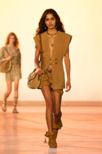 Female model walks a runway in a tan utility vest with matching shorts, carrying a tan crossbody bag and wearing tan suede boots.