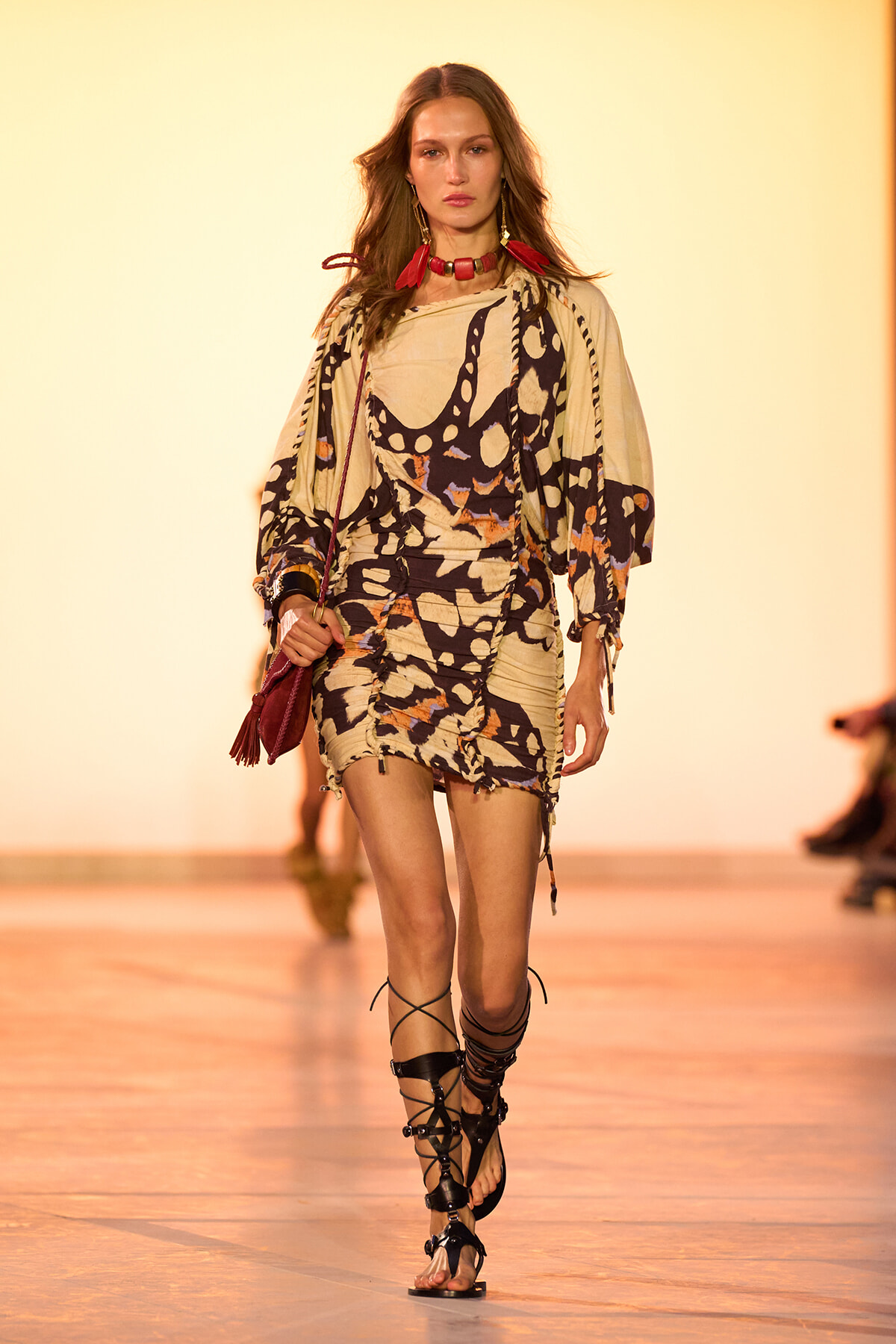 Model walking a runway in a beige and black abstract-print ruched mini dress with billowy sleeves, red statement necklace, and gladiator sandals, carrying a red bag.