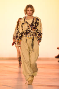 Model walks the runway in a beige utility-style outfit with a bold abstract-print top and wide-leg pants, cinched with a belt and rope details.