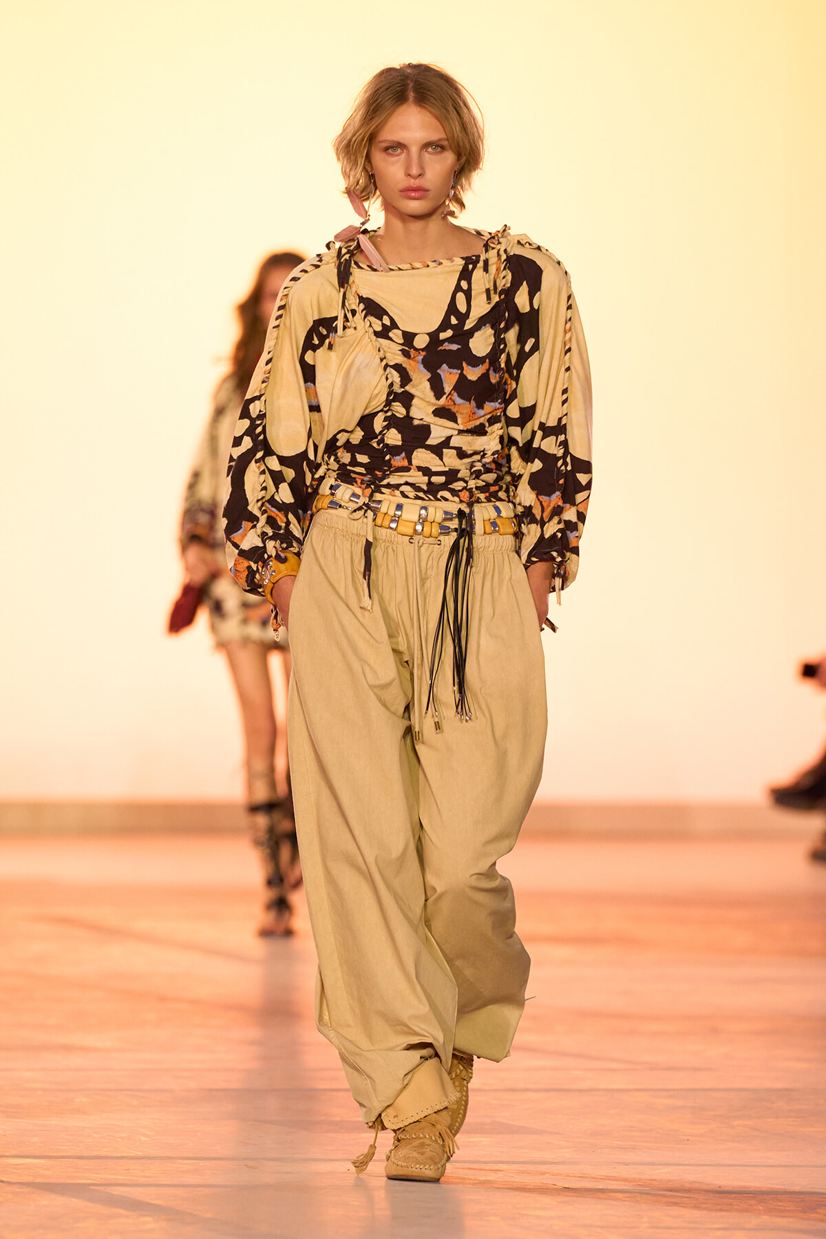 Model walks the runway in a beige utility-style outfit with a bold abstract-print top and wide-leg pants, cinched with a belt and rope details.