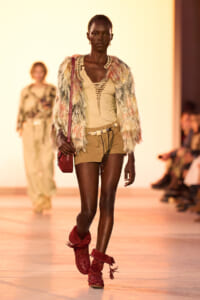 Model walks runway in a multicolored faux fur jacket over a beige lace-up top, tan shorts, red boots, and a burgundy crossbody bag.