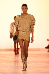 Model walking the runway in a tan oversized top and matching mini skirt, paired with gold gladiator sandals and a small black crossbody bag.
