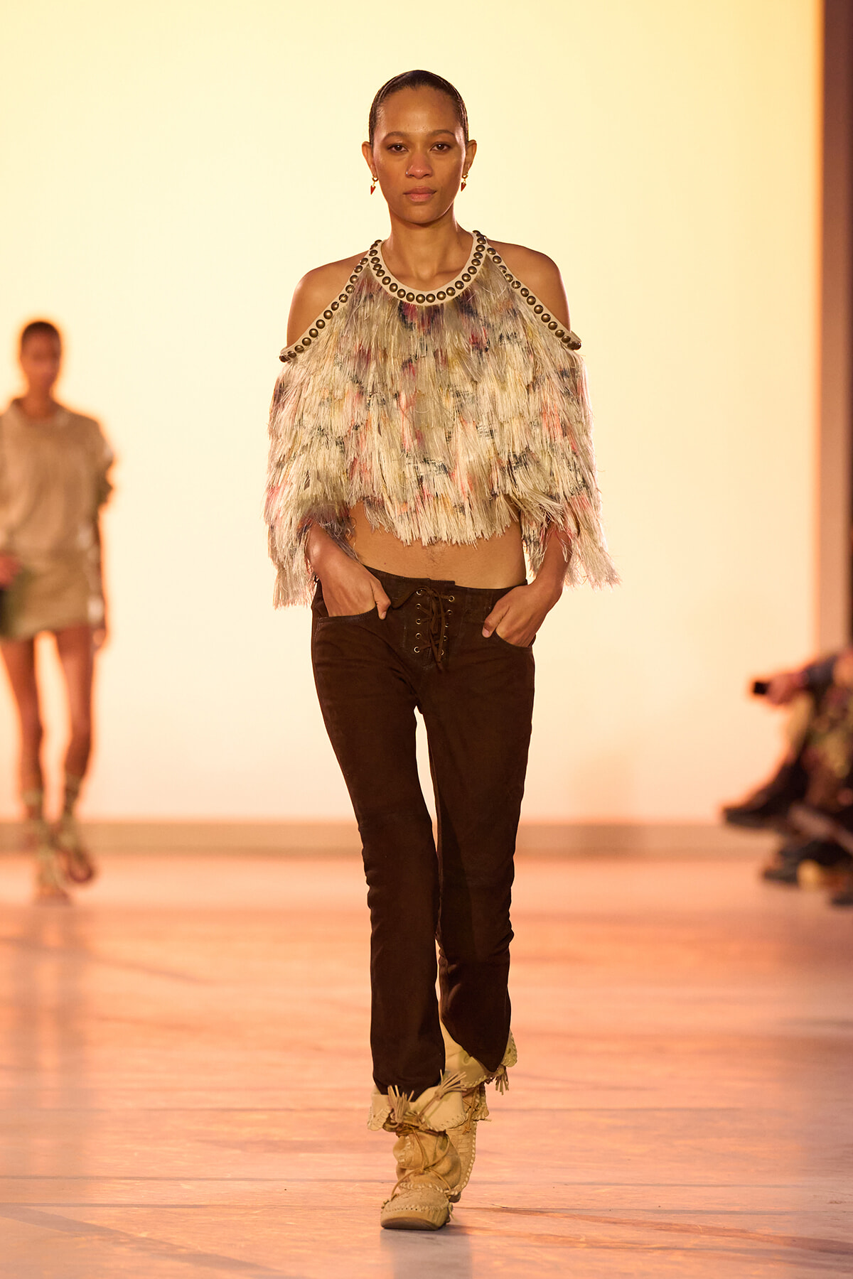 Model walks a fashion runway wearing a multicolored fringe top with metal-studded shoulders, dark brown lace-up pants, and tan boots.