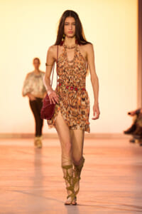 Model walks the runway in a leopard-print halter dress with layered gold jewelry and strappy gladiator sandals, carrying a red bag.