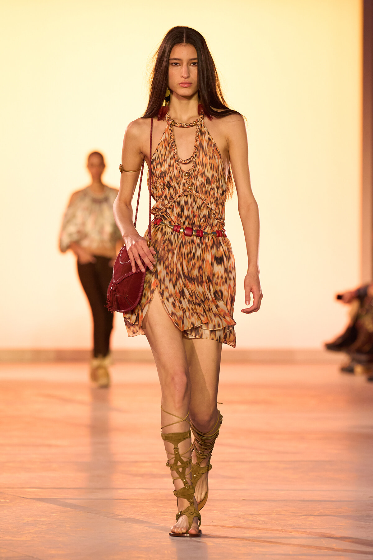 Model walks the runway in a leopard-print halter dress with layered gold jewelry and strappy gladiator sandals, carrying a red bag.