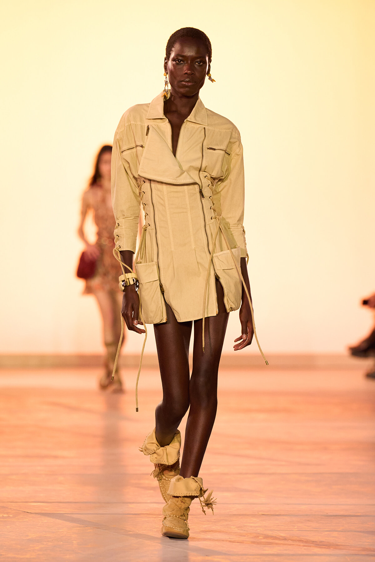 Model walking a fashion runway in a beige utility dress with corset lacing and multiple pockets, wearing matching boots and gold earrings against a warm gradient backdrop.