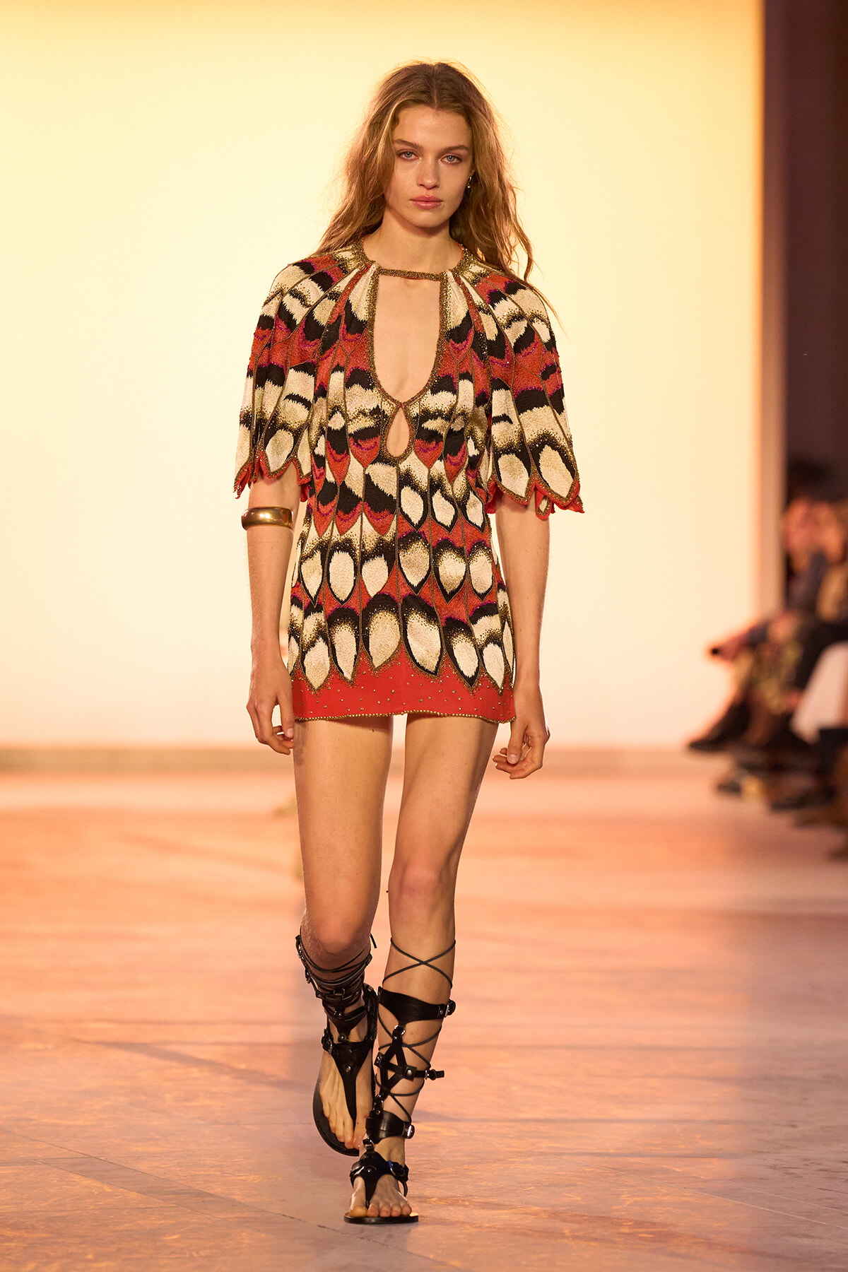 Model on a runway wearing a red, cream, and black patterned mini dress with teardrop cutouts and gold-trimmed sleeves, paired with black gladiator sandals.